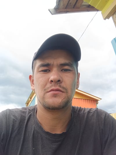 A selfie is taken outdoors, in front of a cloudy sky, with a partially constructed building in the background. A man, wearing a dark t-shirt and a baseball cap, is the central subject. He has short, dark hair and stubble. The building behind him appears to be made of wood and corrugated metal, with sections painted in orange and yellow. The overall scene suggests a rural or developing area.