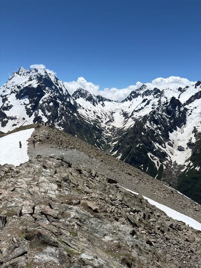 The image depicts a mountainous landscape with snow-capped peaks under a clear blue sky. The foreground features a rocky, barren ridge, with patches of snow scattered on its surface. Two individuals are visible on the ridge; one is standing on a patch of snow further up the ridge, and another appears to be walking in the distance. The midground showcases a vast valley with steep, snow-covered slopes and dark, rocky outcrops. The background is dominated by a range of jagged, snow-covered mountains under a bright blue sky with scattered white clouds. The scene suggests a remote, high-altitude environment suitable for hiking or mountaineering. The general location is inferred to be within the Верхне-Тебердинское сельское поселение, Russia, a region known for its mountainous terrain and natural beauty.