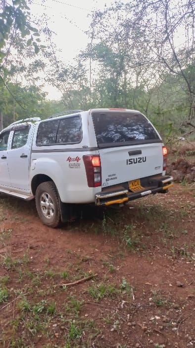 A white Isuzu D-Max pickup truck is parked on a dirt path surrounded by vegetation and trees. The truck has a canopy over the bed and features a "4x4 D-Max" decal. The license plate number KCV 757T is visible. The lighting is soft, indicating either early morning or late afternoon. The surroundings suggest a rural or off-road environment.