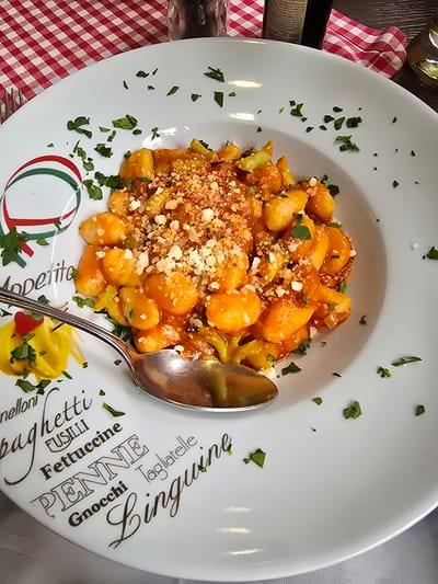 A white ceramic plate, decorated with Italian pasta names and an Italian flag-colored oval, holds a serving of gnocchi in a reddish-orange sauce, topped with grated cheese and chopped parsley. A silver-colored metal spoon rests within the dish. A small yellow condiment with red specks and parsley is visible on the left rim of the plate. The scene is set on a table covered by a red and white checkered tablecloth in Münster, Germany. Partially visible background objects include a silver fork on the left, a clear glass pepper grinder, and a dark bottle labeled "250 mle," likely containing olive oil or balsamic vinegar, on the right. No individuals or active interactions are depicted.