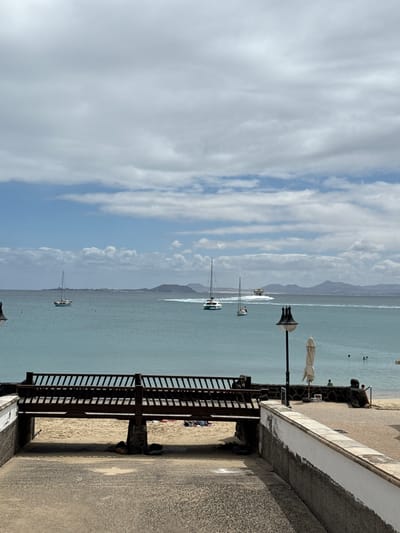 The image is a landscape view from a raised walkway in Playa Blanca, Spain, overlooking a beach and ocean. A light-colored stone or concrete walkway is visible in the foreground, bordered by a weathered wall with a dark trim. A dark wooden railing or fence runs along the edge of the walkway, providing a barrier to the beach below. The beach is sandy and leads to the ocean, which is filled with several sailboats and other watercraft. In the distance, mountainous land is visible. The sky is predominantly blue, filled with scattered white clouds. Several people can be seen on the beach and in the water, suggesting a public recreation area. The overall scene depicts a sunny day at the beach in a coastal town.