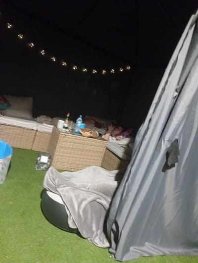 The image depicts a nighttime scene inside a temporary outdoor shelter, possibly a gazebo or tent, set up on a lawn. String lights are visible, casting a warm glow against the dark interior. In the background, two people are lying down on cushioned seating. One person appears to be looking at a phone. A wicker coffee table is in the center of the scene, holding a bottle of beer, a bottle of water, and a glass. To the left, a blue bin is partially visible. In the foreground, a large gray beanbag chair is covered with a blanket. The overall setting suggests a relaxed, outdoor evening gathering. The image was captured in Kobyłka, Poland.