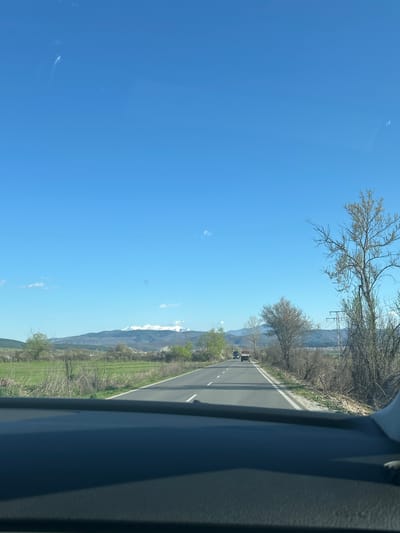 The image depicts a road scene in Hvostiane, Bulgaria, taken from inside a moving vehicle. The view shows a straight stretch of road with two lanes, marked by a white dividing line. There are two vehicles in the distance on the road. To the right of the road is a grassy area, transitioning into scrubland and sparse trees. Beyond this is a mountain range with snow-capped peaks. The sky is clear and blue, with very few clouds. Bare trees branch out near the top of the frame. The edge of the car’s interior is visible on the right side of the image.