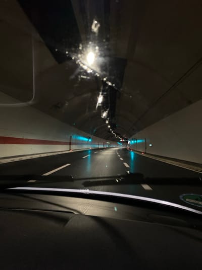 The media file captures an interior view from a moving vehicle, likely a train, as it traverses a dark, artificially lit tunnel. The perspective looks forward and slightly to the right, showcasing the tunnel's infrastructure.
On the left side of the image, the curved concrete wall of the tunnel is visible, reflecting various bright, circular white lights. These reflections appear on what seems to be a window, potentially blurred by speed or condensation, creating a shimmering effect.
Towards the center and extending into the distance, a long, narrow white pathway or platform is prominent, marked with a continuous red stripe down its middle. This pathway is illuminated by evenly spaced blue-green lights embedded along its edge, guiding the way through the darkness.
To the right, a darker, flat surface resembling a track or road runs parallel to the white pathway. This surface features intermittent white dashed lines, similar to road markings, and is also punctuated by more of the blue-green lights. The rightmost edge of the frame shows a dark, textured surface, possibly part of the train's interior or another structure within the tunnel. A green and white sticker or sign is partially visible on the upper right, but its text is illegible.
Further into the tunnel, the lights continue, diminishing in size and intensity with distance, with additional red and green signal lights observable. The environment is entirely dependent on artificial illumination, suggesting it's either nighttime or deep within an underground passage. The scene conveys a sense of forward motion through a highly structured and engineered environment. Given the location context of Pelev Brijeg, Montenegro, the tunnel is most likely a railway tunnel, part of the Belgrade–Bar railway.