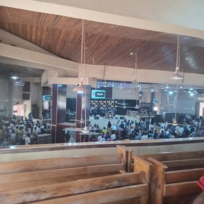 The image shows the interior of a large building, possibly a church or mosque, filled with people seated or kneeling in rows, suggesting a religious service or gathering. The location is specified as Jama A, Nigeria. The building has a high ceiling with wooden paneling and pendant lights. There are wooden pews visible in the foreground. A screen is visible in the center of the room, possibly displaying information related to the service. The people are dressed in various attire, suggesting a diverse congregation. There are podiums where people are standing.