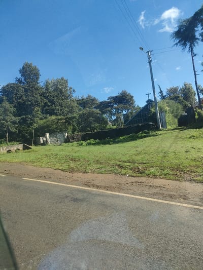 The image shows a rural roadside scene in Kenya, potentially within the Municipality ward. A paved road runs horizontally at the bottom of the frame. A grassy slope rises on the right side of the road. A structure with a triangular, glass-faced façade and a cross on top, likely a small church or chapel, is visible amidst trees and foliage on the hillside. Further to the left, a gated entrance and low stone structure appear in the landscape. A utility pole stands near the chapel. The sky is blue with some clouds. The scene is bathed in daylight.