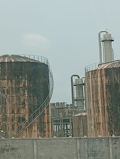 The scene depicts an industrial facility located in Peyer, Nigeria. Two large, cylindrical storage tanks, appearing heavily weathered and discolored with dark brown, black, and orange staining, dominate the midground. The tank on the left features an external spiral staircase ascending to its domed roof, which shows traces of vegetation. Between and behind these prominent tanks, a complex arrangement of industrial infrastructure is visible, including multiple interconnected pipes, metal frameworks, and smaller cylindrical structures. A tall, slender, light-colored industrial tower, capped with an L-shaped pipe, stands towards the right of the frame's center. A concrete barrier wall, light gray and showing some staining, extends across the lower foreground. Small, light-colored objects or blocks are visible at intervals along the top edge of this wall. The sky above is uniformly overcast and pale gray. No individuals or active processes are discernible.