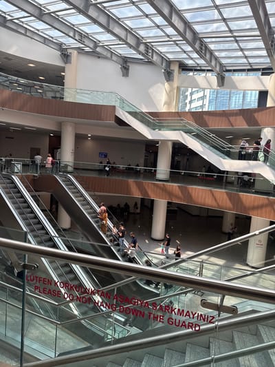 An interior shot captures a modern, multi-level atrium with a glass ceiling and steel beams. Several escalators and staircases, framed by glass and metal railings, connect the different floors. People are seen using the escalators, walking, and standing in various areas. Red text on a glass railing reads "LÜTFEN KORKULUKTAN AŞAĞIYA SARKMAYINIZ PLEASE DO NOT HANG DOWN THE GUARD." The scene suggests a public building, possibly a hospital or a large office complex, located in Çankaya, Turkey, given the Turkish text.
