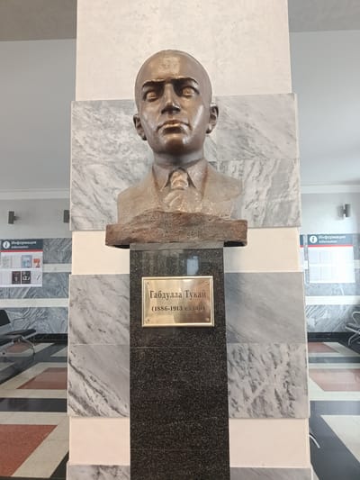 A bronze bust of Gabdulla Tukai, a Tatar poet and writer, is displayed on a pedestal in a well-lit interior space. The bust depicts Tukai from the chest up, with a serious expression. A plaque on the pedestal reads "Габдулла Тукай (1886-1913 еллар)", indicating his name and lifespan in Tatar. The pedestal is made of dark, speckled stone, topped with sections of white and gray marble. Behind the bust, a marble-covered column with gray and white veining serves as a backdrop. To the left and right of the column are informational posters with the heading "Информация Information". The floor in front of the pedestal has a geometric pattern of black, white, and red tiles. The environment appears to be a public building, possibly a museum or cultural center, located in Arsk, Russia. The lighting is bright and even, illuminating the subject clearly. No other people are visible in the frame.