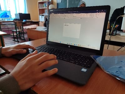 The scene is an indoor computer lab or classroom located in Rishtan, Uzbekistan. In the foreground, a person's hands, with a grey sleeve visible on the left forearm, are positioned over the keyboard and trackpad of an open black HP laptop. The laptop screen displays a Microsoft Word document with a text box containing the partially typed word "RISHTAN." The Windows taskbar, showing various application icons, is visible at the bottom of the screen. The laptop rests on a light brown wooden desk. To the right of the laptop, a blue plastic folder or document protector lies on the desk.

The background reveals a classroom environment. Multiple wooden desks with green-framed chairs are arranged throughout the room. On a desk in the mid-ground, to the left, a computer monitor displays the Windows 11 logo. Further back, another person wearing a light head covering is partially visible behind a computer setup. Another individual is partially obscured on the far right, also at a computer station. A black cylindrical holder containing colored pens or pencils sits on a desk in the mid-ground. A shelving unit filled with books and various items is visible against the back wall, which also features blue curtains over windows and a partial view of a chalkboard.