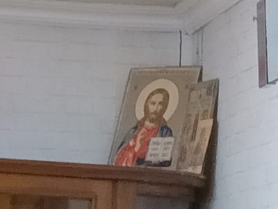The image shows a framed religious icon of Jesus Christ, depicted with a halo and holding an open book. The icon is placed on a wooden shelf, which appears to be part of a larger piece of furniture, possibly a cabinet or a hutch. The background consists of a light-colored wall with horizontal textured lines. To the right of the Jesus icon, another framed object, possibly another icon or a picture, is partially visible. The scene suggests an indoor setting, likely a home or a place of worship. No specific location within Tatysh, Russia, can be identified from the visual information provided.