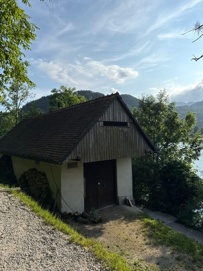 The image shows a small, wooden shed with a brown, shingled roof located in the village of Bled, Slovenia, near Lake Bled (coordinates: 46.363, 14.09). The shed is situated on a gravel path, angled slightly downhill. To the left of the shed, stacked logs form a small pile against the exterior wall. The shed's exterior is painted white with wood paneling on the upper portion.  A single, large wooden door makes up the front facade. The shed has a small, rectangular window, with a view of the lush, forested slopes of the surrounding hills.