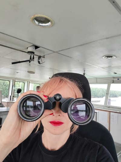 A woman with short, pink hair and bright red lipstick holds a pair of black binoculars up to her eyes. She is wearing a black t-shirt and is seated in what appears to be the pilot's cabin of a vessel. The cabin has large windows that look out onto a body of water and a wooded shoreline, indicating a location along the river system near Kurdiug, Russia. The ceiling of the cabin is white and features overhead lighting fixtures and what appears to be a ventilation system. Behind the woman, a black headrest is visible, and in the distance, through the windows, another person can be faintly seen. The overall impression is one of observation or navigation from within a ship's control room.