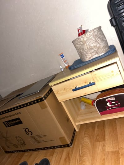 The image depicts a cluttered bedside table and a large cardboard box on the floor. The bedside table is made of light-colored wood and has a drawer with two blue handles. On top of the table are a cream-colored lampshade with a floral pattern, a small tube of sunscreen, a small bottle, and a few flags. Inside the open drawer, there is a yellow can, a clear plastic cap, and a brown and red bag with the words "BOXE ACADEMY" on it.

The cardboard box is labeled "cossette Combinée CAVO" and features the logo "BeBelissimo." The box appears to contain a baby stroller or similar item, as indicated by the illustrations on its side. The floor is made of light-colored wood planks.

The scene suggests a domestic environment, possibly a bedroom, with items in disarray. There are no people visible in the image. The lighting appears to be artificial. The location of Chantepie, France, is not discernible from the visual information in the image.