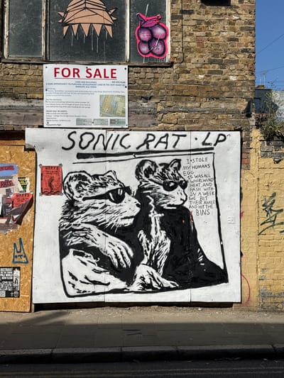 The image captures a scene in Margate, Thanet, United Kingdom. A "FOR SALE" sign advertising "development land in the very heart of Margate Old Town" is posted next to a large piece of street art displaying the words "SONIC RAT LP" and depicting two rats in black and white. The sign lists potential uses for the land including a shop, basement, holiday flats, parking, and a pool. Underneath the artwork, various smaller posters and flyers are pasted. The wall the artwork is on is adjacent to a brick wall with graffiti. The sidewalk runs alongside the art installation.