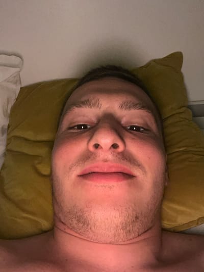 A close-up image shows a person lying down, with their head and upper chest filling the frame. The image is oriented with the person's head on the left side, as if looking upwards from a prone position. The subject has short, dark hair and visible stubble or light beard growth on their chin and upper lip. Their dark eyes are open and looking towards the camera, and a soft, contented smile plays on their lips.
The person's head rests on a soft, crumpled, greenish-yellow (olive-colored) pillow. The lighting is dim and warm, creating a relaxed atmosphere, possibly suggesting evening or nighttime. To the left of the person's head, a plain, light-colored wall or headboard is visible, while a white sheet or blanket can be seen beneath the pillow on the bottom right. The scene appears to be indoors, likely in a bedroom. There is no visible text in the image. The general location context is Vitry-sur-Seine, France.