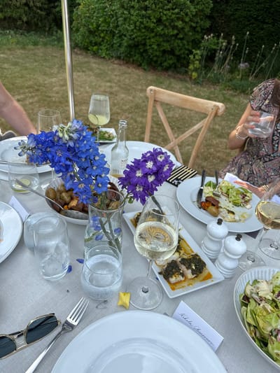 A partly outdoor dining scene is captured from a high, slightly angled perspective. A white tablecloth covers the table on which various dishes, glasses, and flowers are arranged. Several plates of food including salads and what appears to be fish are visible. White salt and pepper shakers sit next to the food plates. Wine glasses, some filled with white wine, are also present. Vases holding blue and purple delphinium flowers create a center piece. A name card reading "Nikola Simeonow" rests near a white plate. Sunglasses and silverware lie on the table. A woman wearing a patterned dress holds a glass in the upper left corner. In the background is a green lawn with plants, suggesting an outdoor garden setting. Based on the style of table setting and the surrounding greenery, it is highly likely to be in the city of Cambridge, United Kingdom.