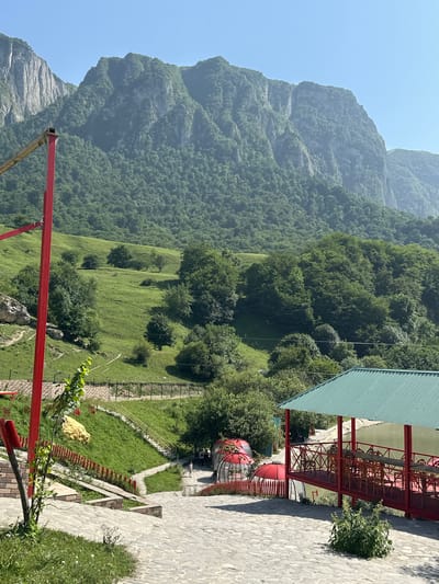 The image depicts a view from an elevated paved area looking towards a verdant mountainside. In the foreground, a red structure, possibly a playground element, is visible on the left. To the right, a covered seating area with a green roof and red railings overlooks a body of water. Further down a grassy slope, several red dome-shaped structures are present. A stone pathway with yellow railings leads down the slope. The background is dominated by a large, tree-covered mountain range under a clear blue sky. The scene appears to be a recreational area in a mountainous region, possibly Qırızdəhnə, Azerbaijan, given the context. There are a few indistinct figures visible near the red dome structures.
