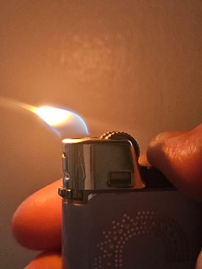 The image shows a close-up, eye-level, indoor shot of a person's hand operating a purple disposable lighter with a flame. The lighter has a metallic grinding wheel and a purple body with a circular dotted pattern. The flame is bright orange and blue and appears to be burning steadily. The background is a warm, out-of-focus brown color. The image appears to be taken in the city of Mäntsälä, Finland.
