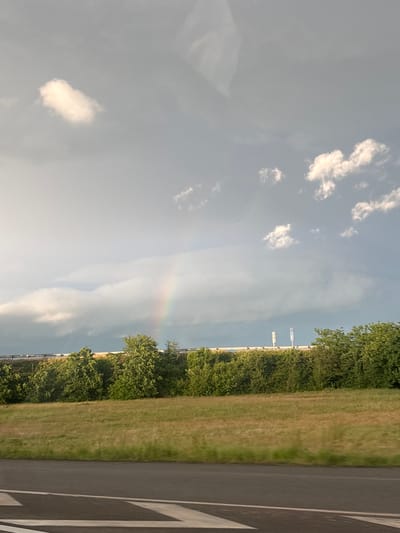 A landscape photograph, rotated 90 degrees counter-clockwise, captures a sky with scattered white clouds, a faint rainbow arc in the middle of the image, and a thin line of white structures (potentially a bridge) near the horizon. A band of green vegetation (trees and grass) borders the right edge, adjacent to a paved surface with painted chevron markings. The location is Châtenois, France.