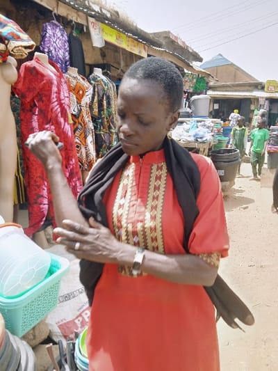 The image portrays an active outdoor market scene in Kadoma, Zimbabwe, under bright daylight. A woman of African descent, appearing middle-aged, stands prominently in the foreground. She has short, dark hair and is dressed in a reddish-orange tunic featuring intricate gold and black embroidery on the chest and collar, with a black scarf draped over her shoulders. A watch with a black strap is on her left wrist, and a silver-toned ring adorns her left ring finger. Her head is tilted downwards, and she is observed touching her left forearm with her right hand, which also holds a small, dark cylindrical object.

To the immediate left and behind the woman, a clothing stall is visible, displaying various garments on mannequins and hangers, including items with red tie-dye, purple tie-dye, and geometric patterns. In the mid-ground and background, the market extends with unpaved ground and multiple stalls featuring corrugated iron roofs. Various goods are displayed, including light blue/green plastic baskets, colorful plastic buckets, and large sacks, one of which is marked "INDONESI PP." Several other individuals, including two children dressed in green shirts, are visible further in the background, moving through the market area.
