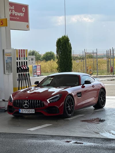 A red Mercedes-Benz AMG GT is parked at a Shell V-Power gas station in Drazhevo, Bulgaria. The car is positioned at a gas pump, with the front grille and headlights clearly visible. The license plate reads "CB 9708 KH". In the background, there are gas pumps, a tall green tree, a chain-link fence, and a field of dry grass. The sky is partly cloudy. The ground around the car appears wet, with puddles reflecting the vehicle and the surrounding environment.