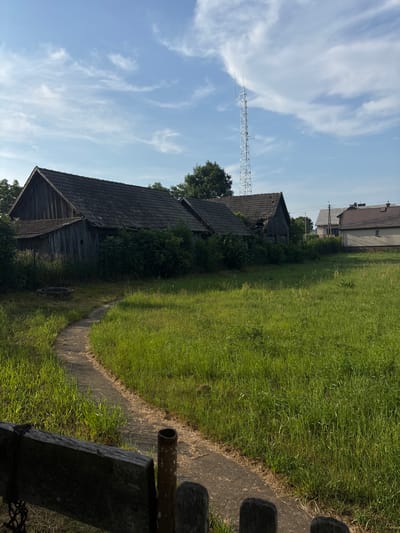 The image depicts an outdoor scene on a sunny day in Janów, Poland. In the foreground, a wooden bench and a metal fence post are visible, partially obscuring a concrete pathway that curves to the left. The pathway leads through a field of tall, green grass.
Further back, on the left side of the image, are several dilapidated wooden outbuildings with dark, weathered roofs. These structures appear old and somewhat neglected, with overgrown bushes surrounding them. To the right of these outbuildings, in the middle ground, is a tall, metallic communication tower. Beyond the outbuildings and the tower, several houses with tile roofs are visible, some appearing more modern and well-maintained than the older structures in the foreground.
The sky is blue with scattered white clouds, and the sun casts distinct shadows, indicating it is likely midday or early afternoon. The overall impression is a rural or semi-rural setting, with a mix of old and new buildings, and a natural, overgrown landscape. There are no people visible in the image.