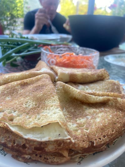 The image captures a close-up view of a plate of folded crepes, likely part of a meal. In the background, a person is visible, out of focus, eating with a fork. Also in the background, out of focus, are a bowl of shredded carrots and a bunch of green onions. The setting appears to be outdoors, with greenery and natural light suggesting a garden or patio area, possibly in Fadeyevo, Russia. The crepes are golden brown and appear to be thin and delicate.