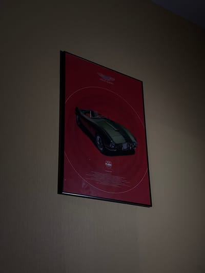 A framed poster depicting a dark green Aston Martin classic convertible is displayed on a light brown, vertically textured wall. The poster features a prominent red background with concentric white circles and the "ASTON MARTIN" winged logo in the upper center. A smaller, circular logo, potentially "RETRO CLASSICS," is visible in the lower left corner above lines of fine print text. The poster is encased in a simple black frame. The scene is dimly lit, suggesting an indoor environment, with the lighting creating subtle reflections on the poster's surface. No people, discernible actions, or further environmental details are visible. The location, specified as Lebyazh'e, Russia, cannot be determined from the visual evidence in the image.