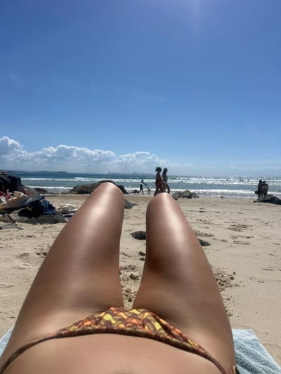 A first-person, medium shot captures a sunbather reclining on a beach in Coolangatta, Australia. The scene is oriented vertically. The individual's legs, tanned and seemingly oiled, dominate the foreground. Part of a patterned bikini bottom is visible. The beach, a sandy expanse, is speckled with people in the distance. The ocean, reflecting sunlight, is visible between the legs of the sunbather. A blue sky with scattered clouds occupies a substantial portion of the frame.