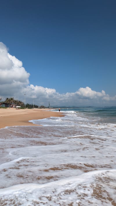 This image displays a daytime coastal scene on a sandy beach in Thành phố Tuy Hòa, Vietnam. In the immediate foreground, white seafoam from receding waves covers wet, light brown sand. The midground features a wide expanse of dry, light brown sand leading towards a line of palm trees and other green foliage. One individual, wearing dark shorts, is observed walking alone at the water's edge, facing right. Multiple tall, white utility poles are spaced along the back of the beach, and several white beach umbrellas or shelters are visible among the trees on the left. In the distance on the right, industrial structures, possibly cranes, are discernible near the coastline. The background consists of a blue-green ocean with small waves, meeting a horizon where distant mountains or hills are visible under a blue sky with scattered white cumulus clouds.