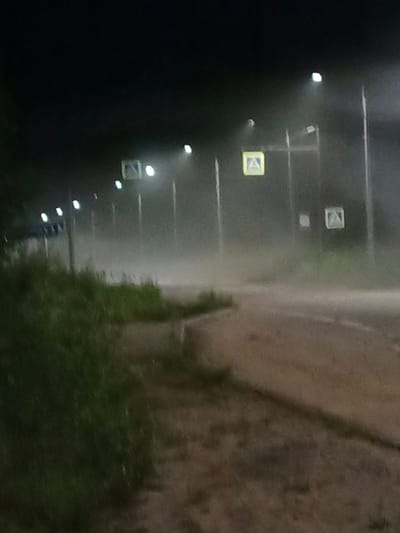 The image depicts a nighttime scene on a road with a pedestrian crossing. There are streetlights illuminating the area, creating a foggy or misty atmosphere. Several yellow pedestrian crossing signs are visible along the roadside. The road surface appears to be asphalt. To the left, there is a grassy area with some vegetation. The overall impression is one of a somewhat hazy and dimly lit urban or suburban environment.