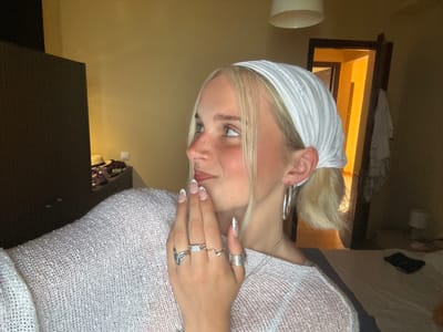 A young woman with blonde hair, wearing a white headscarf and a white mesh sweater, is pictured in what appears to be a bedroom. She is wearing several rings on her fingers and a nose ring. Her nails are manicured with white tips and small gems. The lighting suggests it is daytime, possibly late afternoon, given the warm glow emanating from a window. The setting includes a dark piece of furniture that could be a cabinet or closet, and a white cylindrical object in the foreground. The woman's face is turned to the side, and her expression is relaxed and somewhat pensive. The image is oriented sideways. There is no visible text.