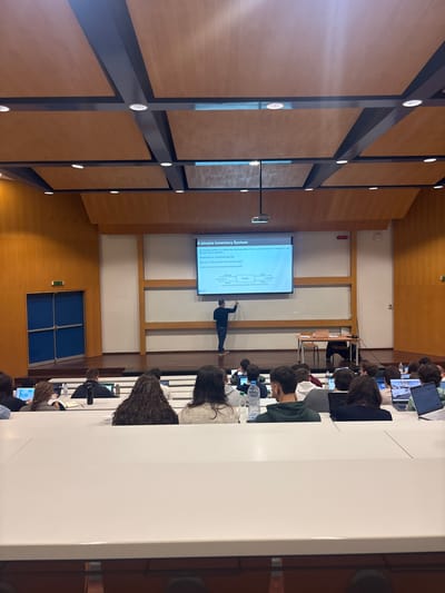 The image depicts an indoor lecture hall or large classroom in Porto Salvo, Portugal. A male lecturer, dressed in a dark blue shirt and dark pants, stands at the front of the room on a raised wooden stage. He is facing a large white whiteboard, holding a marker in his right hand, and appears to be writing or pointing towards it. Above the whiteboard, a projector screen displays a slide titled "A simple Inventory System," featuring text and a flowchart diagram. A projector is mounted from the ceiling, casting the image onto the screen.

In front of the stage, approximately two dozen students are seated in tiered rows of white desks and dark chairs. The students are primarily visible from their backs and heads. Several students are using open laptops, some are observing the front, and others have notebooks or water bottles on their desks. The room features light wood-paneled walls with darker trim and horizontal white sections. A set of double blue doors is visible on the far left wall. The ceiling consists of a grid of rectangular wooden panels with integrated circular lights and dark structural beams. The overall environment suggests an academic or professional presentation setting.