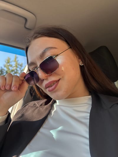 A woman with long brown hair is angled sideways in what appears to be the interior of a car. She is wearing sunglasses, a white top, and a black blazer. Her left hand holds the sunglasses near her face. A portion of a blue sky and tree canopies are visible outside the car window. The location is not discernible as Zaosinovo, Russia based on this image alone.