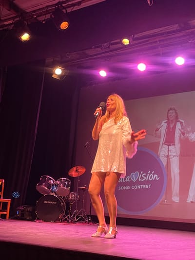 A female singer in a sparkly, one-shoulder mini-dress is performing on a stage. She is holding a microphone and has her left arm extended. The stage is lit with pink and purple lights. To the left of the stage, a drum set is visible. Behind the singer, a screen displays an image of two men in white suits and red shirts, with the text "Eurovisión SONG CONTEST" and "16-Save your kisses for me" visible. The background appears to be a dark curtain. The overall scene suggests an indoor performance, likely a concert or competition. The presence of the text "Eurovisión SONG CONTEST" indicates the event is related to the Eurovision Song Contest. The text "Ibersound" is visible on the bass drum. The singer is wearing sparkly heeled shoes.