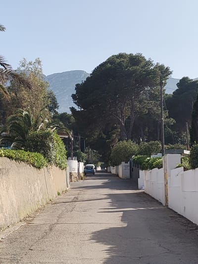 The image depicts a narrow, slightly curving street in Rotes, Spain. The road surface is asphalt, showing signs of wear and tear with cracks and patching. The street is bordered on both sides by walls, one a textured, beige-colored wall and the other a white wall. Occasional landscaping and trees are visible over these walls. In the distance, a blue car is traveling away from the viewer. Behind the car and street scene, a mountain range is visible under a clear, blue sky. Utility poles run along the street's edge.