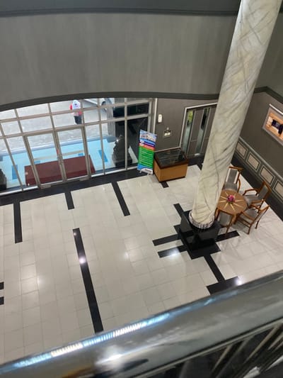 This is an overhead forensic view of the ground floor interior of a building, identified as being in Cipeundeuy, Indonesia. The floor is composed of polished white tiles interspersed with black rectangular patterns. A large, cylindrical pillar with white and grey marble veining rises from a black square base on the right side of the frame. Adjacent to this pillar, a round wooden table with a small pink floral arrangement is surrounded by four light-upholstered chairs.
At the rear, a large glass double-door entrance with silver frames allows natural light. A person wearing a white headscarf and a red top is partially visible through the glass, appearing to stand or walk outside. A red mat lies on the floor directly inside the entrance. To the right of the entrance, a dark-topped wooden console is positioned next to a vertical banner displaying blue and green sections with text, partially revealing 'BANKU' at its base. An obscured dark, possibly sculptural, object is visible through the glass on the left side of the entrance. Grey walls define the space, with decorative wainscoting visible on the right-hand wall. A reflective metal railing occupies the lower foreground, indicating the elevated viewpoint.
