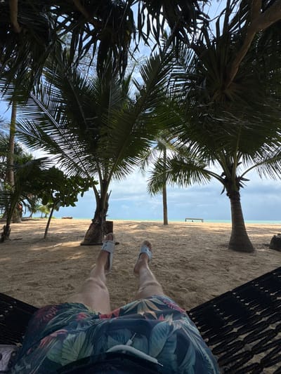 The image depicts a person's legs and lower torso, clad in floral-patterned shorts and light-colored slide sandals, lying in a black hammock. The hammock is positioned on a sandy beach, with the person's feet facing away from the viewer. The background reveals a tropical beach scene. Several palm trees with lush green fronds cast shadows on the sand, with their trunks visible. The sand is light brown and appears smooth, with some areas showing footprints or indentations from the hammock. Beyond the beach, a calm, turquoise body of water stretches to the horizon, meeting a cloudy sky with patches of blue. A small, dark-colored bench or structure is visible in the distance on the beach, near the water's edge. The overall environment suggests a relaxed vacation setting in Ban Khuk Khak, Thailand.