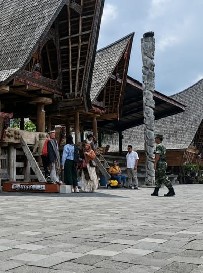 In Ambarita, Indonesia, a group of people are gathered in an open courtyard in front of traditional Batak architecture. The buildings feature distinctive, high-pitched roofs covered in wooden shingles and ornate, dark wood trim. A tall, carved stone totem stands prominently to the right of the frame, depicting a series of human faces stacked vertically.
In the foreground, the ground is paved with large, grey square tiles. Several individuals are interacting or observing the surroundings. On the left, a mannequin dressed in traditional attire stands near a carved wooden object. Two women are standing near the mannequin, one in a blue top and dark pants, the other in an orange headscarf and a patterned dress. Further back, a man in a white shirt and tan pants stands, and a woman in a yellow shirt sits with a yellow bag beside her. To the right, a soldier in camouflage uniform walks away from the camera, his back facing towards the viewer. The overall atmosphere appears to be a cultural or historical site.