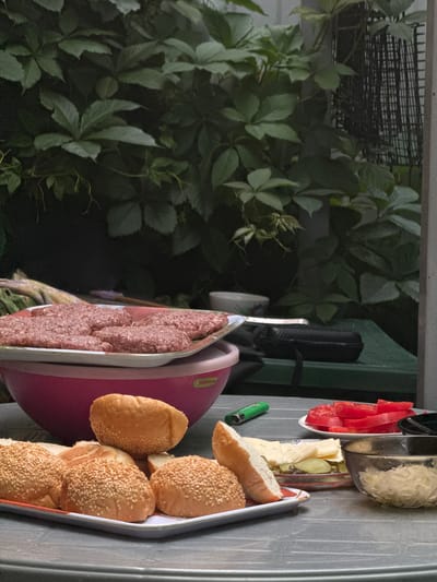 A close-up, eye-level, outdoor shot captures a table set for a barbecue in Pribrezhnyi, Russia. In the foreground, a platter holds several sesame-seed topped burger buns, some halved, revealing their white interiors. Beside this platter are slices of cheese, pickles, and chopped red tomatoes on a smaller plate. To the right, a clear bowl contains shredded cabbage. A green lighter rests on the table near the tomatoes. In the background, a large pink bowl contains raw hamburger patties on a white tray, some of which are overlapping. Behind the table, lush green foliage covers the background, creating a natural, slightly overgrown setting. A dark, zipped pouch is visible on a green surface behind the pink bowl.