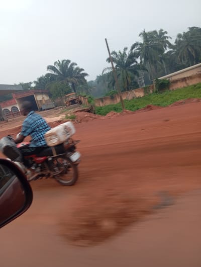 The image captures a scene in Imilike, Nigeria, likely on a cloudy day, depicting a man riding a red motorcycle on a reddish-brown dirt road. The motorcycle is laden with two large, white, rectangular containers strapped to the rear. The rider, seen from the side and slightly behind, is wearing a blue and black checkered shirt. In the background, there are buildings, some with arches and a red roof, and palm trees. A small, weathered yellow vehicle is visible further down the road. The overall impression is one of motion, with the road and surrounding elements blurred due to the movement of the camera.