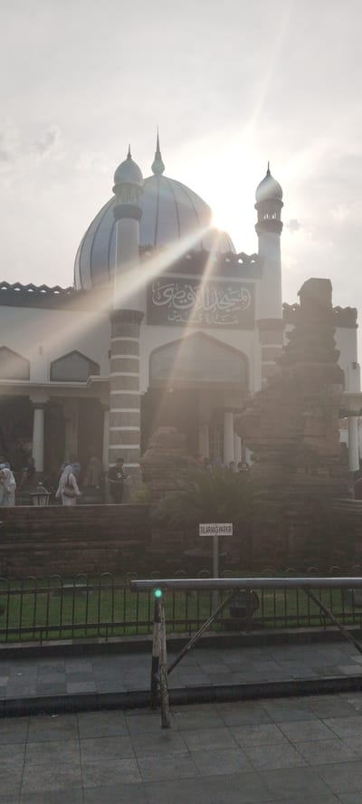 The image shows the exterior of the Great Mosque of Kauman in Yogyakarta, Indonesia, bathed in the bright sunlight of late afternoon, which creates lens flares and overexposure in parts of the image. The mosque features a prominent blue dome and minarets, with intricate architectural details. Arabic calligraphy is visible on the facade above the entrance.

In the foreground, there is a metal fence and a paved walkway. To the right, a weathered brick structure stands, possibly an older part of the mosque complex or a historical artifact. Several people, some wearing headscarves, are seen walking on the stairs leading up to the mosque and in the background. A sign that reads "DILARANG PARKIR" (which translates to "NO PARKING" in Indonesian) is visible on a post near the fence. The sky is bright with some clouds, and the sun is positioned high and slightly to the left of the frame. The overall scene suggests a public space during a time of religious observance or gathering.