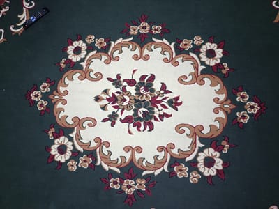 This is a top-down, eye-level, full shot of a dark green rug with a large, intricate floral pattern in the center. The pattern features an oval-shaped cream-colored area with a bouquet of flowers in dark red, beige, and dark green. The floral motif extends outward with swirling brown and cream-colored borders and additional smaller flowers in the same color palette scattered across the dark green background. A black remote control is placed in the upper left corner of the rug, angled diagonally. The lighting appears to be indoor, possibly from overhead sources, creating some subtle shadows. The image is forensic in nature, providing a detailed, objective view of the rug and the remote. The location within the city of Ӯртабуз, Tajikistan, is not ascertainable from the image itself.