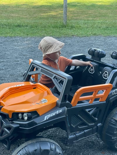 A young child, wearing a tan sun hat and an orange t-shirt, sits in the driver's seat of a black and orange toy electric vehicle. The vehicle has a roll cage, large tires, and the word "SPORTS" is visible on the side. The child is holding onto the steering wheel. The scene appears to be outdoors, with gravel on the ground and a grassy area in the background. The lighting suggests it is daytime, possibly late afternoon due to the warm tones. The image was taken at a slightly downward angle, focusing on the child and the toy car.