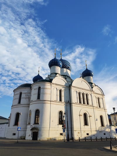 A white church with blue domes topped with golden crosses stands against a bright blue sky filled with white, wispy clouds. The church has multiple domes, arched windows, and ornate architectural details. In the foreground, a paved street and sidewalk are visible, with a black metal fence separating the sidewalk from the street. A lamppost with a traffic light and a pedestrian crossing sign are also present on the sidewalk. The sun casts long shadows, suggesting it is either early morning or late afternoon. The image was taken in the city of Selivanovo, Russia.