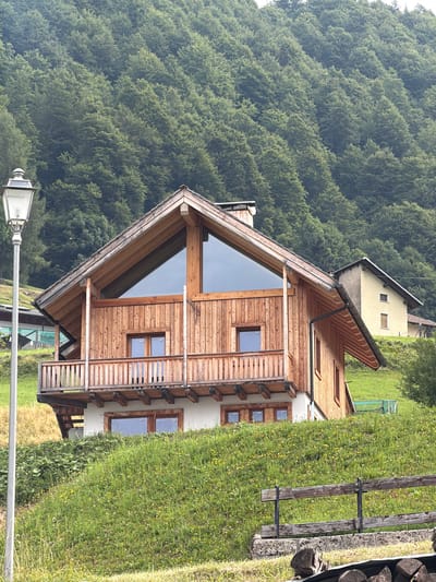 A wooden chalet-style house is situated on a grassy hillside in Sauris / Zahre, Italy. The chalet has a prominent gable end with a large triangular window and a wooden balcony. The exterior walls are clad in vertical wooden planks. In front of the chalet, a lamp post stands on the left side of the frame. The hillside is covered in green grass with scattered wildflowers. Behind the chalet, a dense forest of trees rises up the slope, creating a backdrop of lush greenery. To the right of the chalet, a smaller, yellow building with a pitched roof is visible. A rustic wooden fence runs across the foreground of the image. The scene is set in a mountainous, rural environment, typical of the Carnic Alps region.
