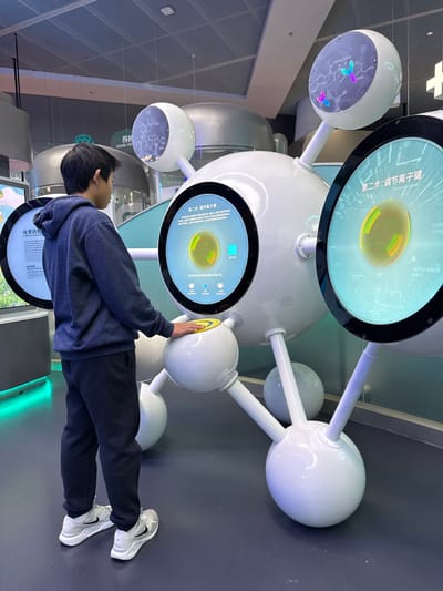 An indoor scene in an exhibition space within Guangming District, China, depicts a young male interacting with a large, white, multi-spherical interactive display. The male, observed in profile facing left, wears a dark blue hooded sweatshirt, dark pants, and white athletic shoes with grey accents, a Nike swoosh visible on his right shoe. His right hand rests on a prominent yellow circular button integrated into a lower sphere of the main display.

The central display is a complex, futuristic-looking structure composed of interconnected white spheres and rods, resembling a molecular model. It features multiple integrated screens. The screen directly in front of the male displays Chinese characters "第二步：调节离子键" (Step 2: Adjust Ionic Bonds) above a graphic of a luminous yellow-green spherical object. An overhead screen shows intricate chemical structural formulas. To the left, a partially visible screen features Chinese text including "化学" (Chemistry) and "CHEMICAL DRUGS."

The environment includes a dark blue-grey floor and a light blue-green partition wall behind the main exhibit. In the background, other exhibition elements are visible, including large, rounded, metallic structures and additional screens. The ceiling is light-colored with a grid pattern and integrated lighting fixtures. A white cross symbol on a white panel is partially visible in the far right background.