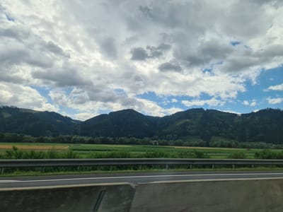 The image shows a view from a vehicle driving on a highway, likely in Kraubath an der Mur, Austria. In the foreground, a concrete barrier and a metal guardrail separate the highway from the adjacent landscape. Beyond the highway, lush green fields and crops stretch towards a backdrop of densely forested mountains. The sky is partly cloudy, with patches of blue sky visible amongst dramatic, gray clouds. The scene depicts a rural, mountainous environment.