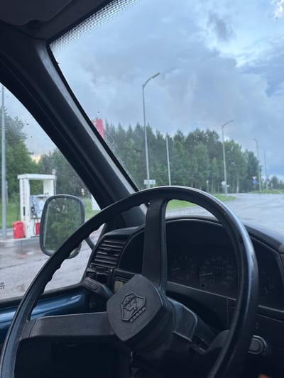 This is a point-of-view shot from inside a car, looking through the windshield. The steering wheel and dashboard are in the foreground. Outside the car, a gas station with a white and red structure is visible on the left. Beyond the gas station, a road curves to the right, lined with trees and streetlights. The sky is overcast with clouds. The image appears to be taken in Chernovskoye, Russia, as indicated by the Russian-made car's dashboard emblem, which resembles that of a Lada. The road conditions suggest it might be raining, as indicated by the water on the windshield.